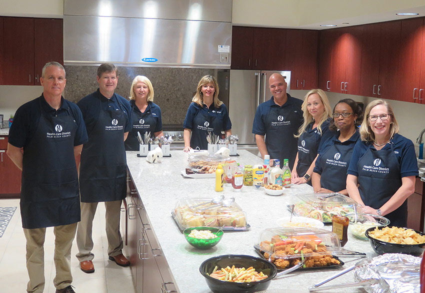Executives wearing Health Care District branded aprons stand around a kitchen island