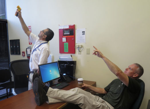 Al aims a temperature reader at a vent. Rick reclines in a chair with feet on the desk and points